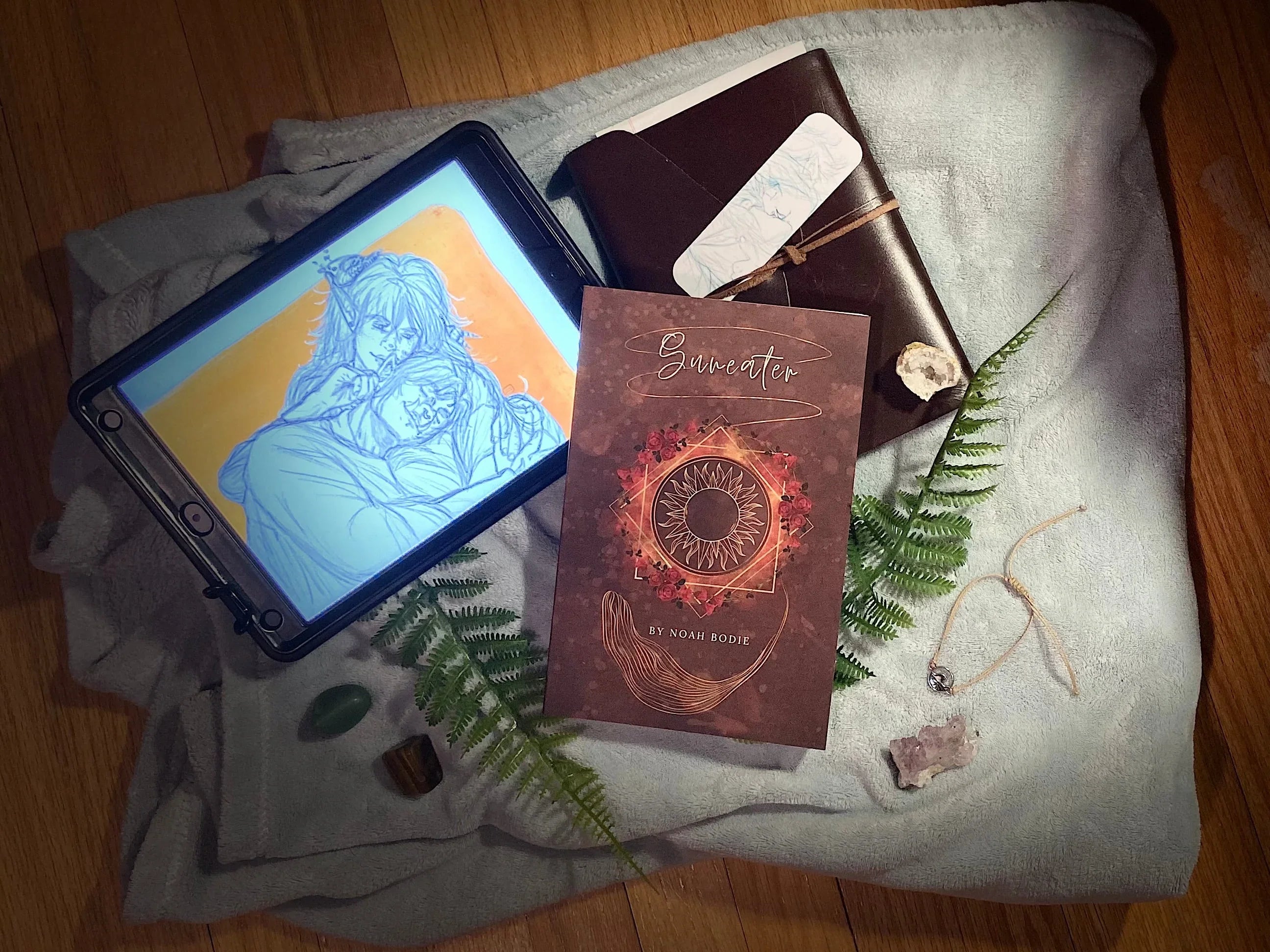 Book titled Suneater by Noah Bodie on cloth with digital drawing on tablet, crystals, fern leaves, leather journal, and necklace