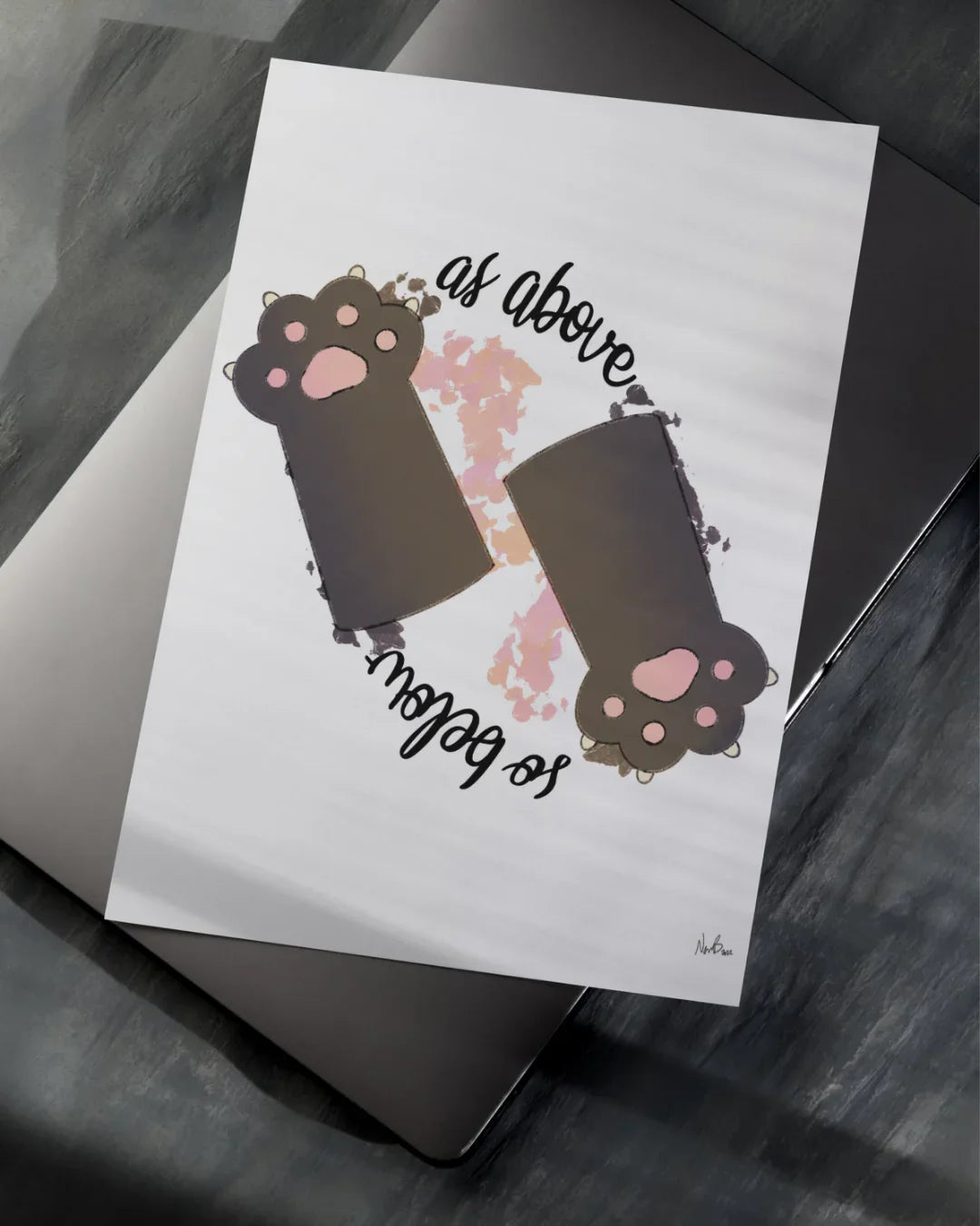Printed artwork with two black cat paws and text 'as above so below' on white paper over laptop