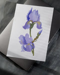 Watercolor painting of purple iris flowers on white paper resting on closed laptop on dark surface